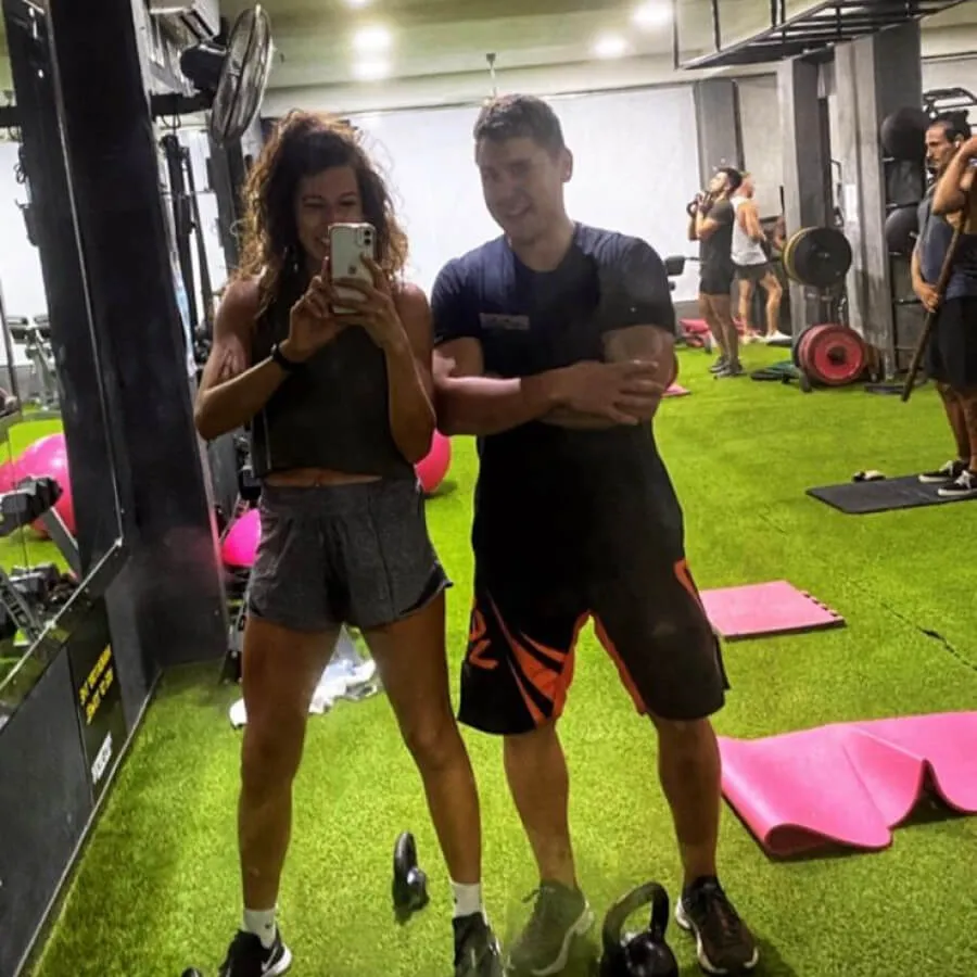 Two people taking a gym mirror selfie on artificial green turf, with gym equipment and other people exercising in the background.