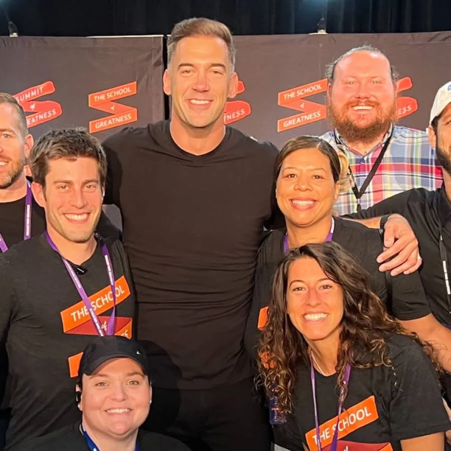 Group of smiling people wearing black t-shirts with orange 'The School' badges posing together.