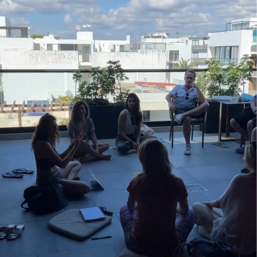 Group of people sitting in a circle on a terrace with a cityscape background under a partly cloudy sky.
