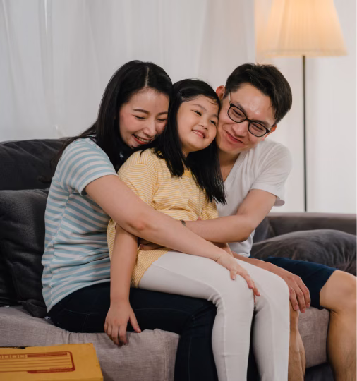 Smiling Asian family of three sitting closely on a couch, mother hugging daughter while father looks on, with a lit floor lamp in the background.
