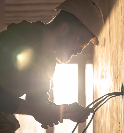 Man wearing a safety helmet working with electrical wires on a wall, with bright sunlight behind him.