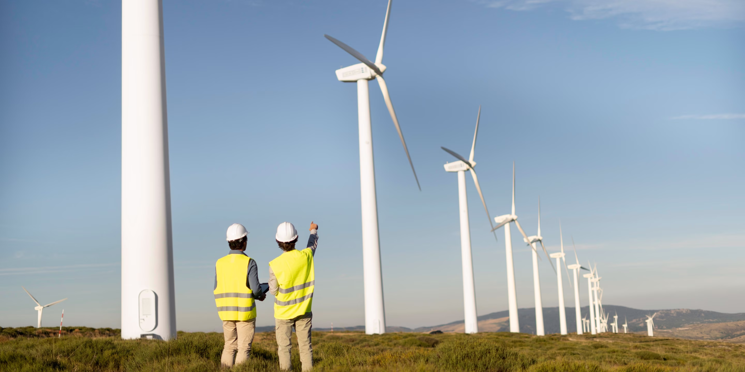 Two workers in yellow safety vests and helmets inspecting a row of wind turbines in a green field under a clear blue sky.