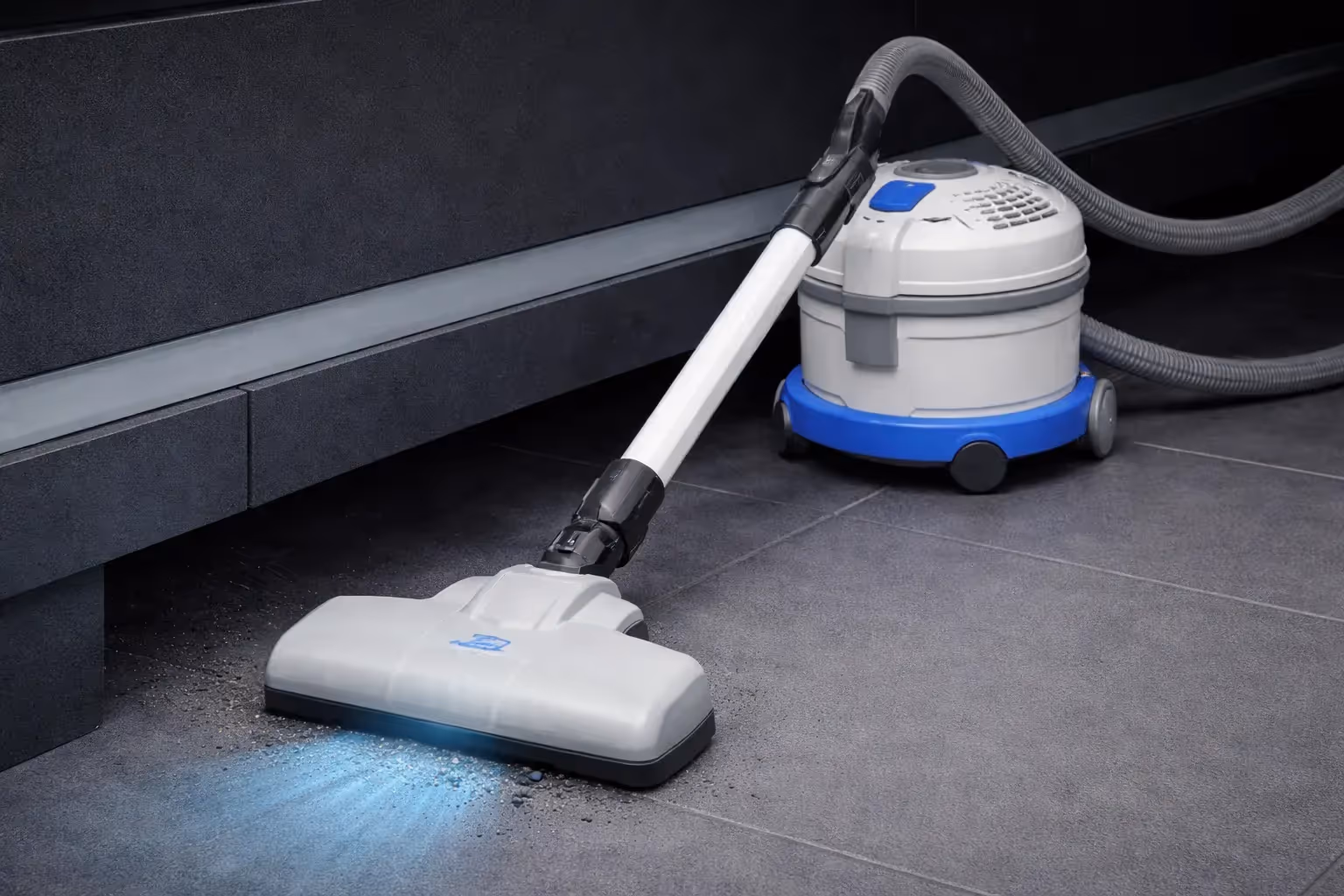 Modern canister vacuum cleaner with blue light suction head cleaning gray tiled floor near dark furniture.