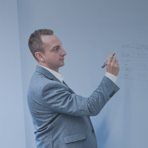 Man in a gray suit writing on a whiteboard with a marker.