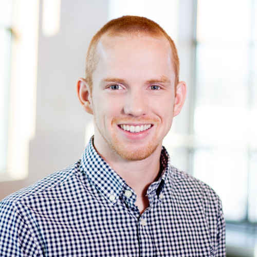 Smiling young man with short red hair wearing a blue and white checkered shirt in a bright office setting.