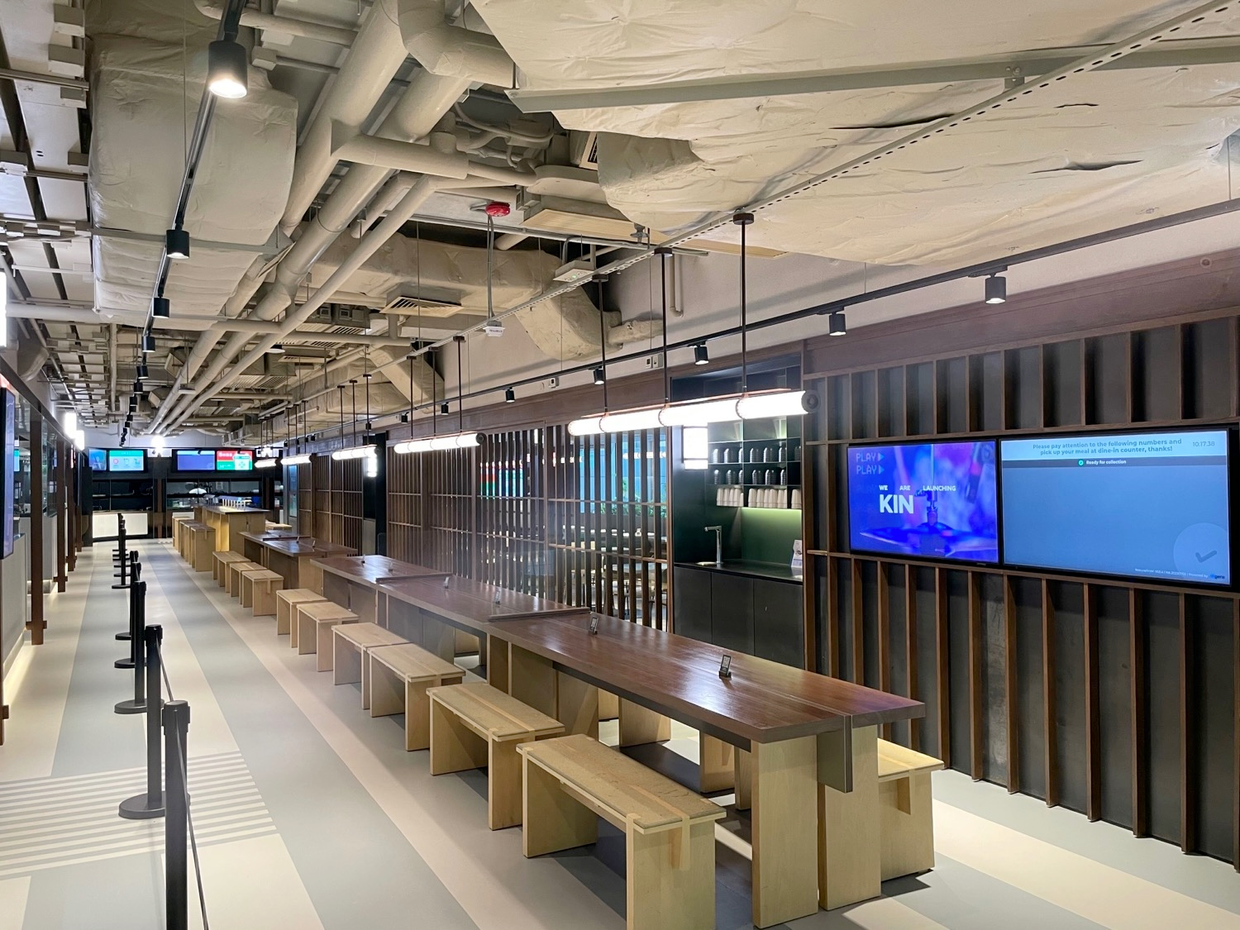 Empty modern dining area with long wooden tables, benches, hanging lights, and digital screens on the wall.