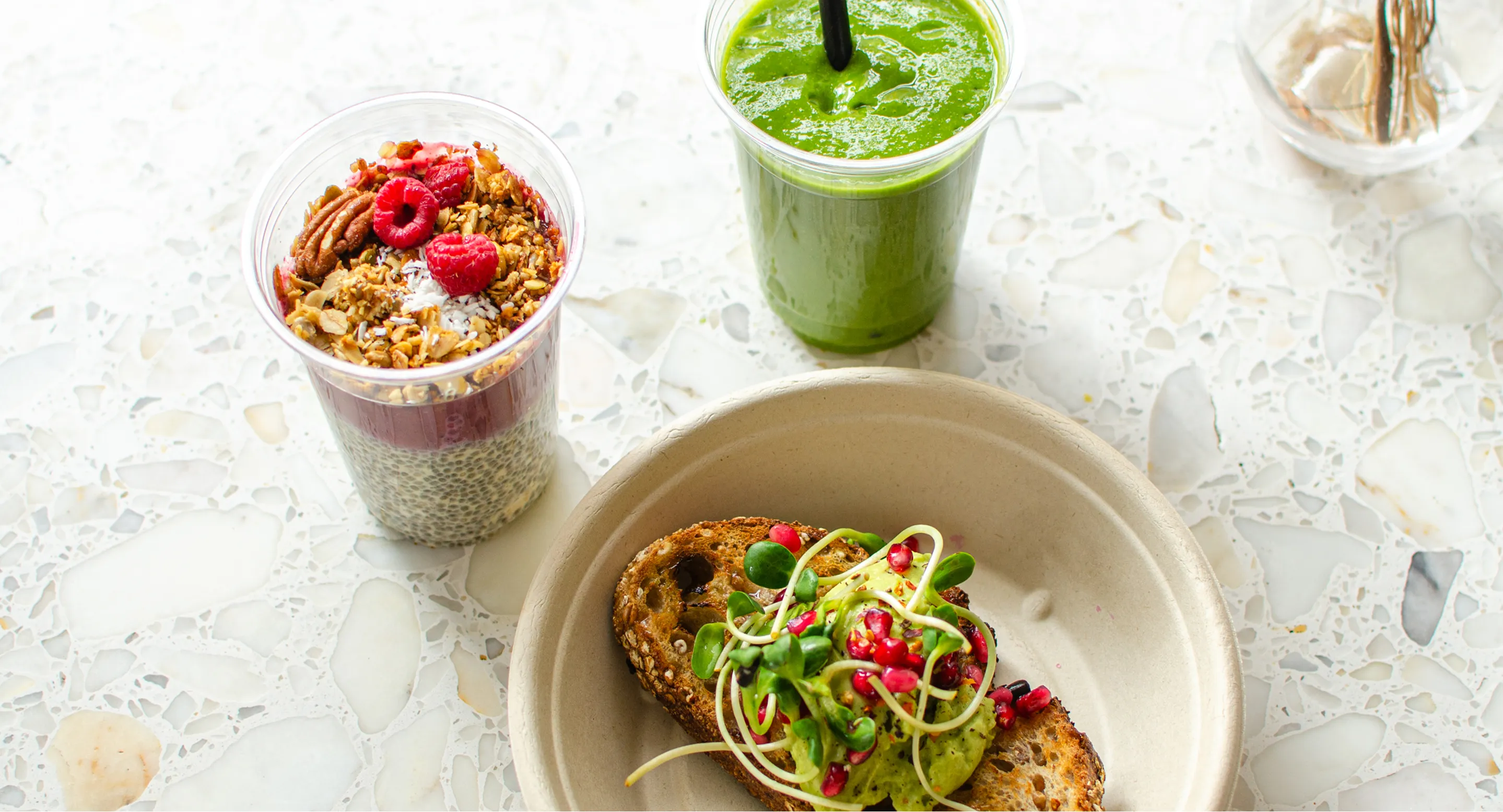 Kulhollinen avokadolla, ituilla ja granaattiomenan siemenillä päällystettyä ruisleipää, vihreä smoothie ja chiavanukas tuoreilla vadelmilla, pähkinöillä ja granolalla kirkkaissa muovimukeissa valkoisella kivipöydällä.
