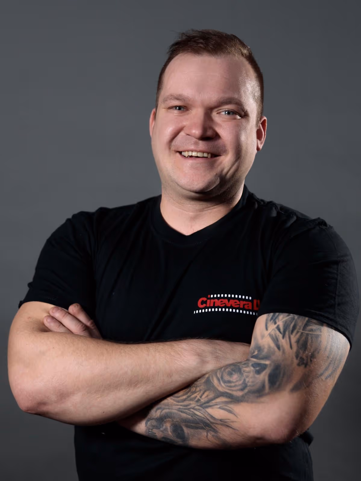 Smiling man with folded arms showing a tattoo sleeve on his right arm wearing a black Cinevera shirt.