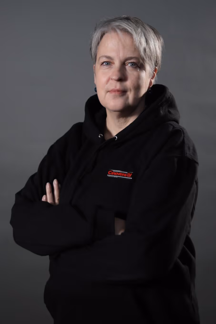 Mature woman with short gray hair standing with arms crossed wearing a black hoodie against gray background.