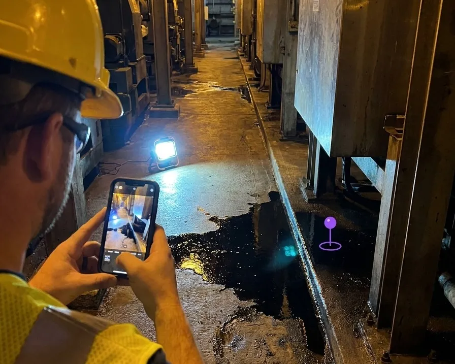 A worker raising a phone toward a spill in an industrial corridor, with a position marker pinned to the floor at the hazard
