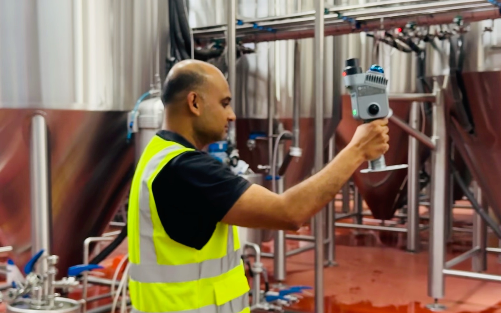 An operator in a high-visibility vest walks a brewery tank farm holding a handheld LiDAR scanner