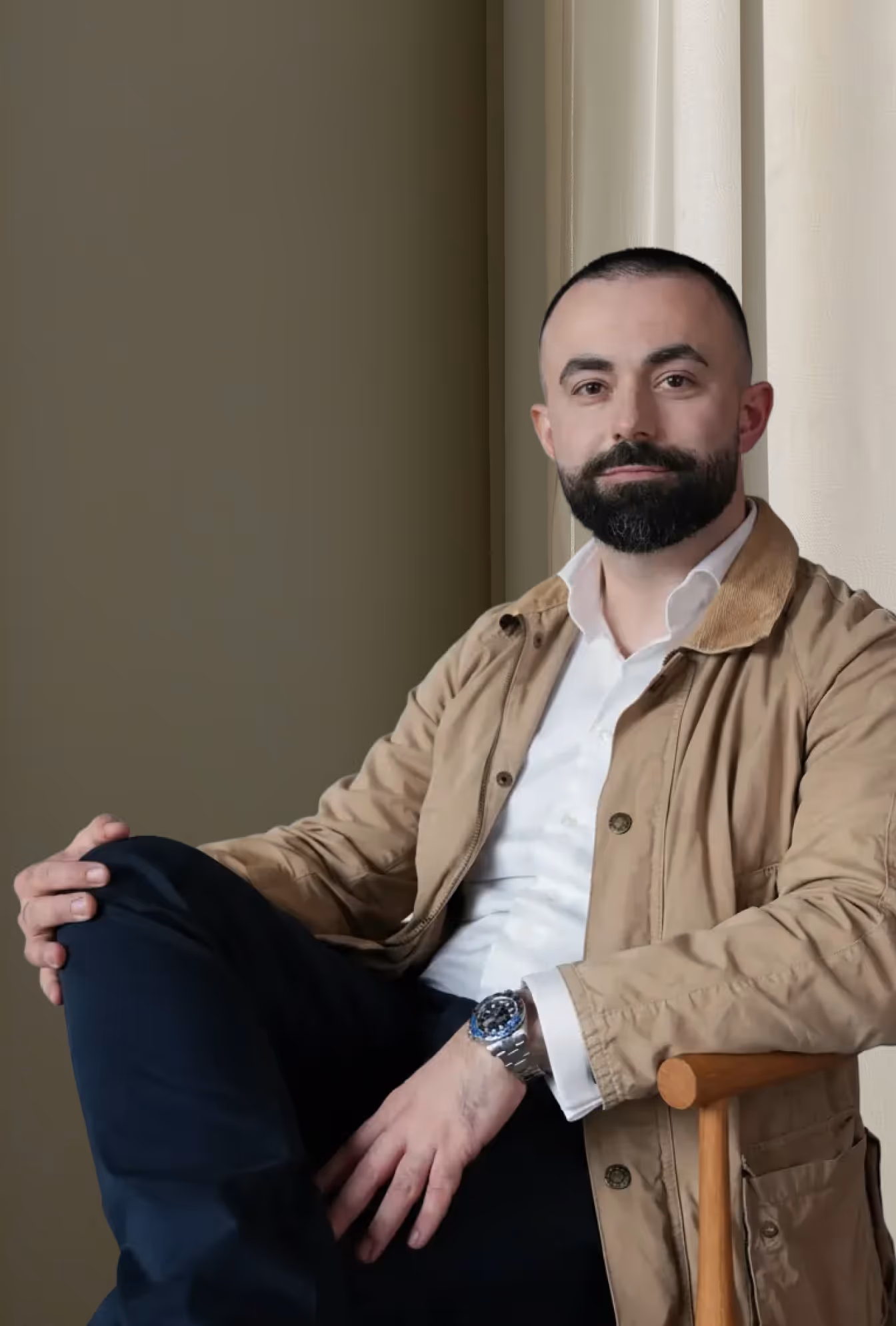 Bearded man seated on wooden chair, wearing a tan jacket, white shirt, and dark pants, posing calmly indoors.