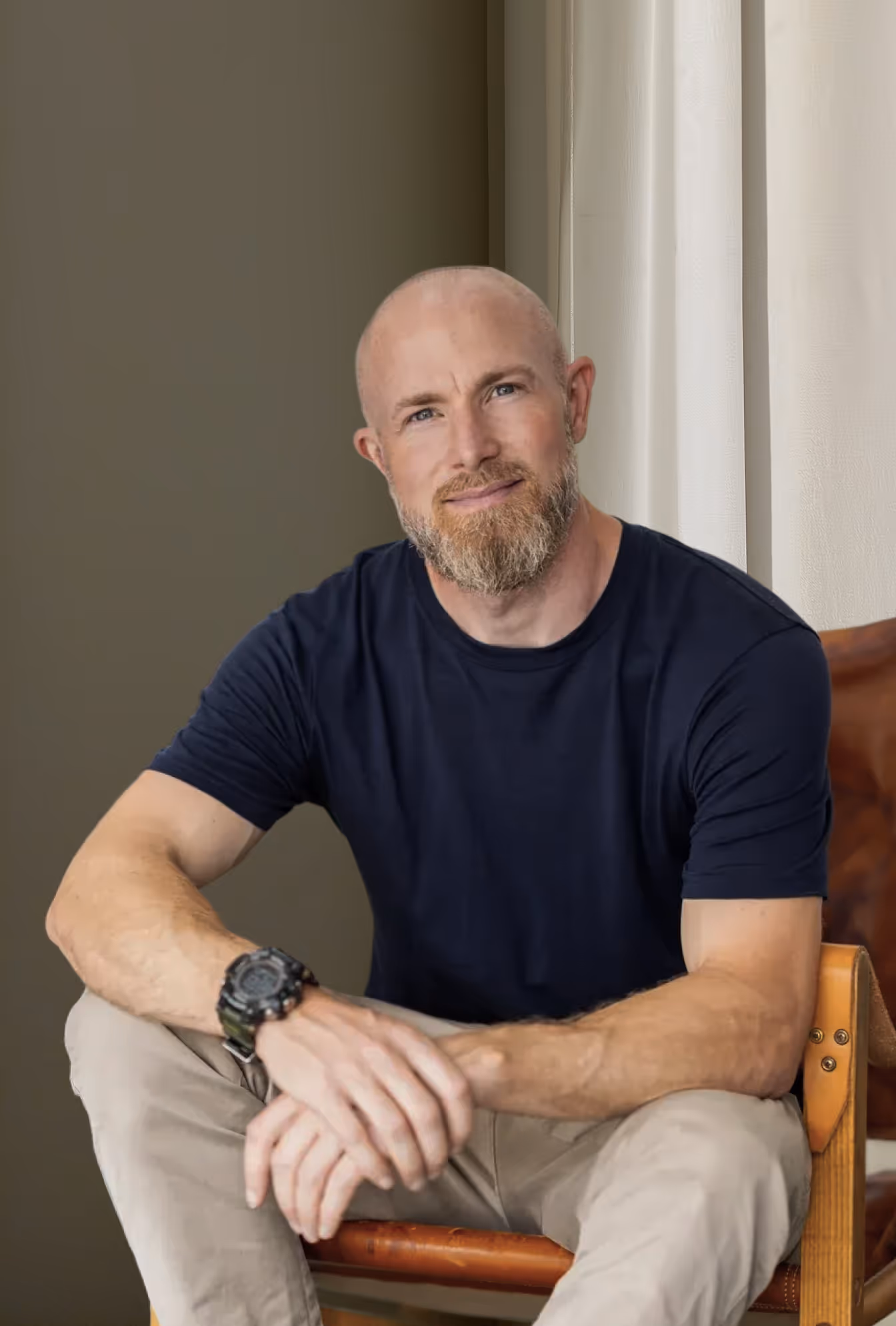 Bald man with a beard wearing a black t-shirt and beige pants sitting on a wooden chair.