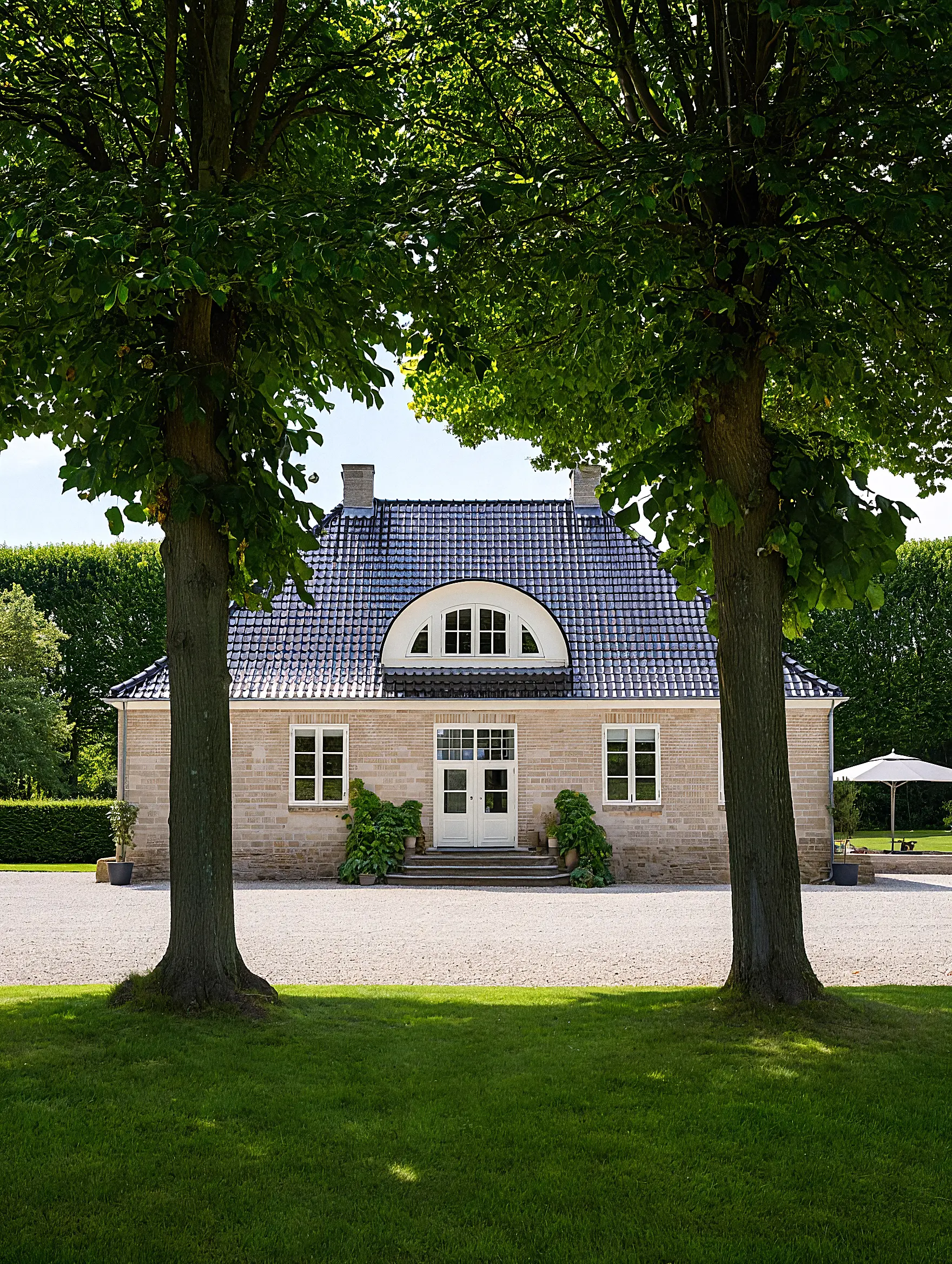A beige brick house with a dark tiled roof framed by two large leafy trees and surrounded by green grass and shrubs.