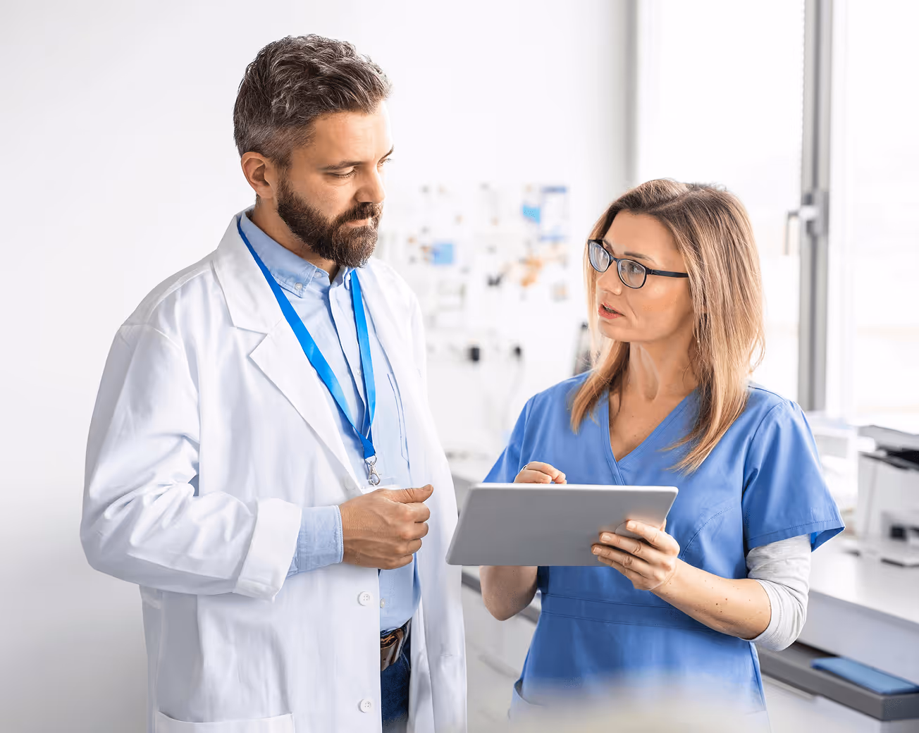 Female nurse in blue scrubs showing a tablet to a male doctor wearing a white coat in a clinical setting.