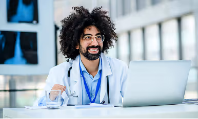 Smiling male doctor with curly hair and glasses sitting at a desk with a laptop in a bright medical office.