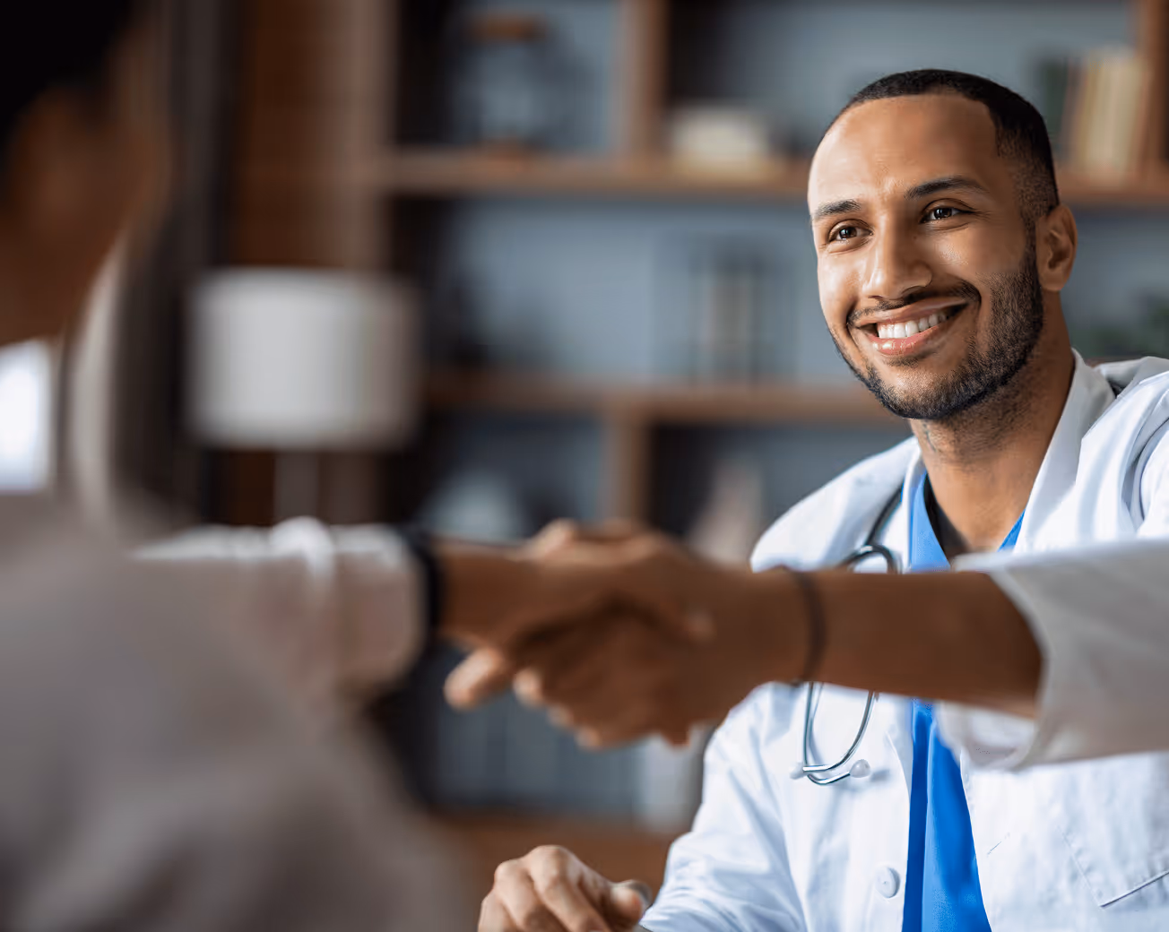 Smiling male doctor wearing a white coat and stethoscope shaking hands with a patient in an office.