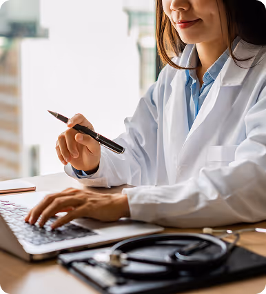 Female doctor wearing a white coat working on a laptop with a stethoscope on the desk.