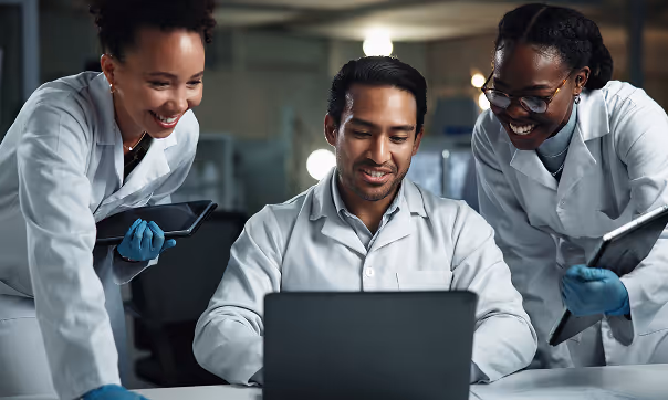Three scientists in white lab coats and blue gloves smiling and looking at a laptop screen in a laboratory.