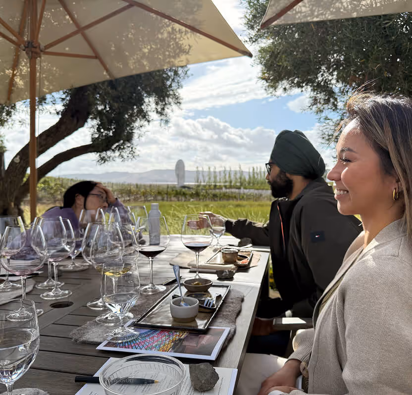Three people seated at an outdoor table with wine glasses and food plates, enjoying a sunny day with umbrellas and trees nearby.