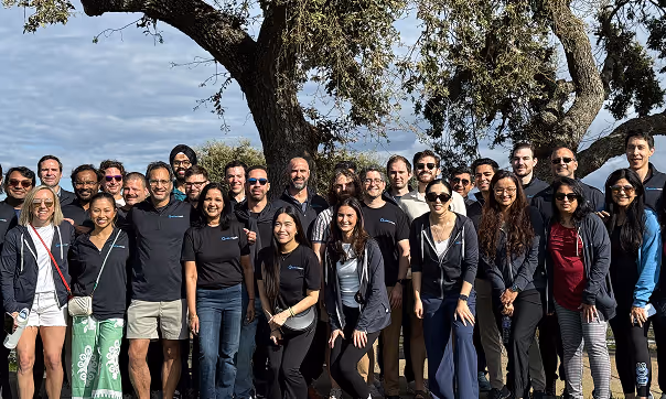 Group photo of a diverse team of 26 people standing outdoors in front of a large tree under a cloudy sky.