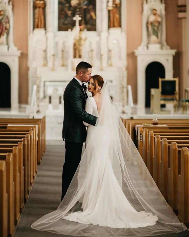 Bride and groom embracing in a church aisle, with the groom kissing the bride's forehead and her veil flowing behind her.