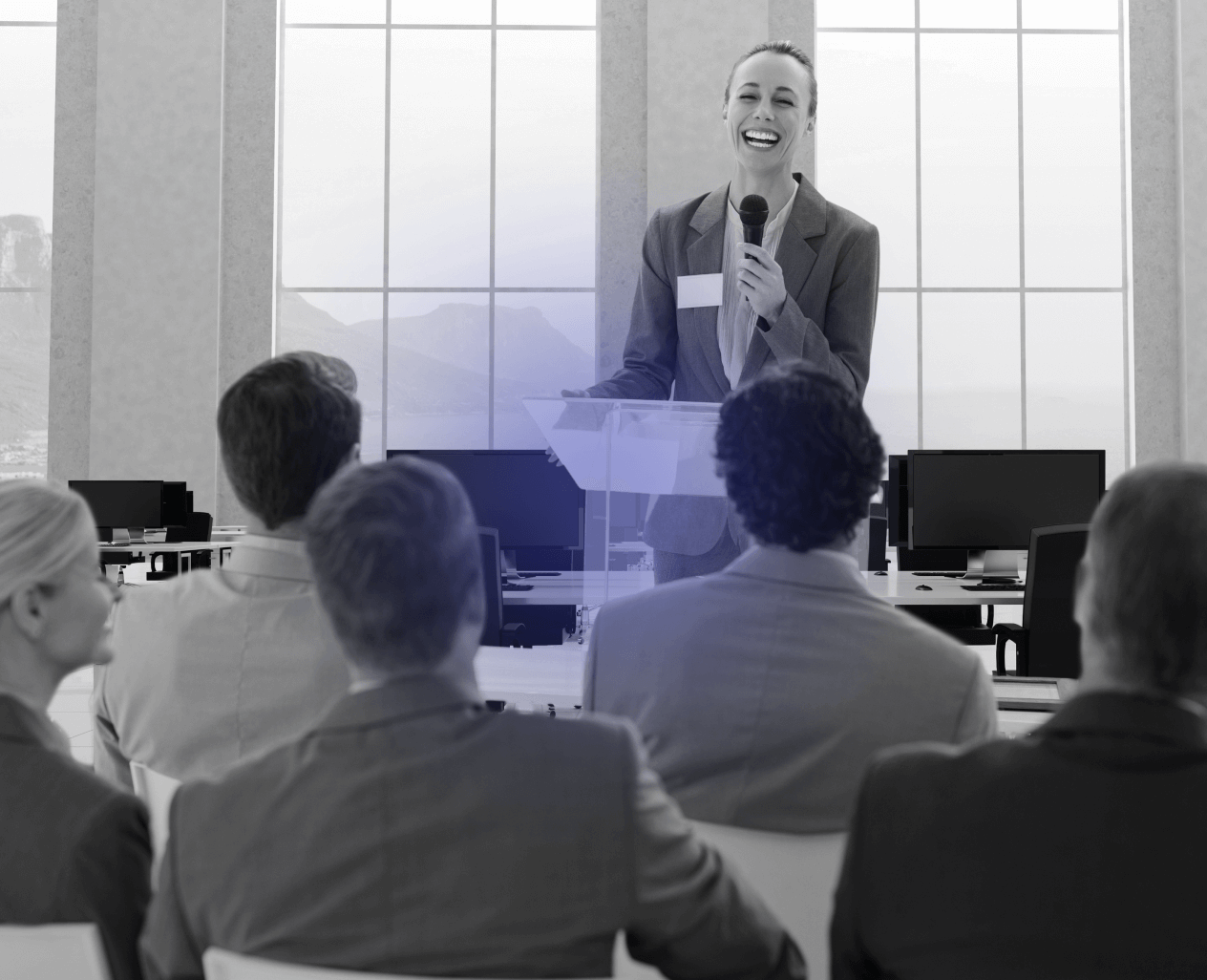 Smiling woman in business attire speaking into a microphone at a podium in an office setting with seated audience.