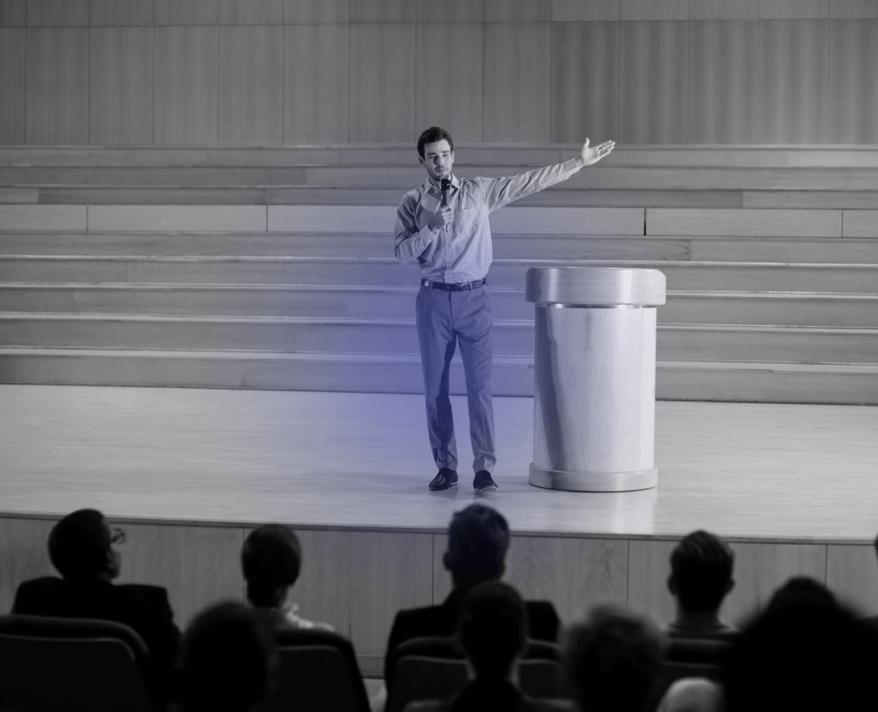 Man standing on stage holding a microphone and gesturing with his arm extended, speaking to an audience seated in front.