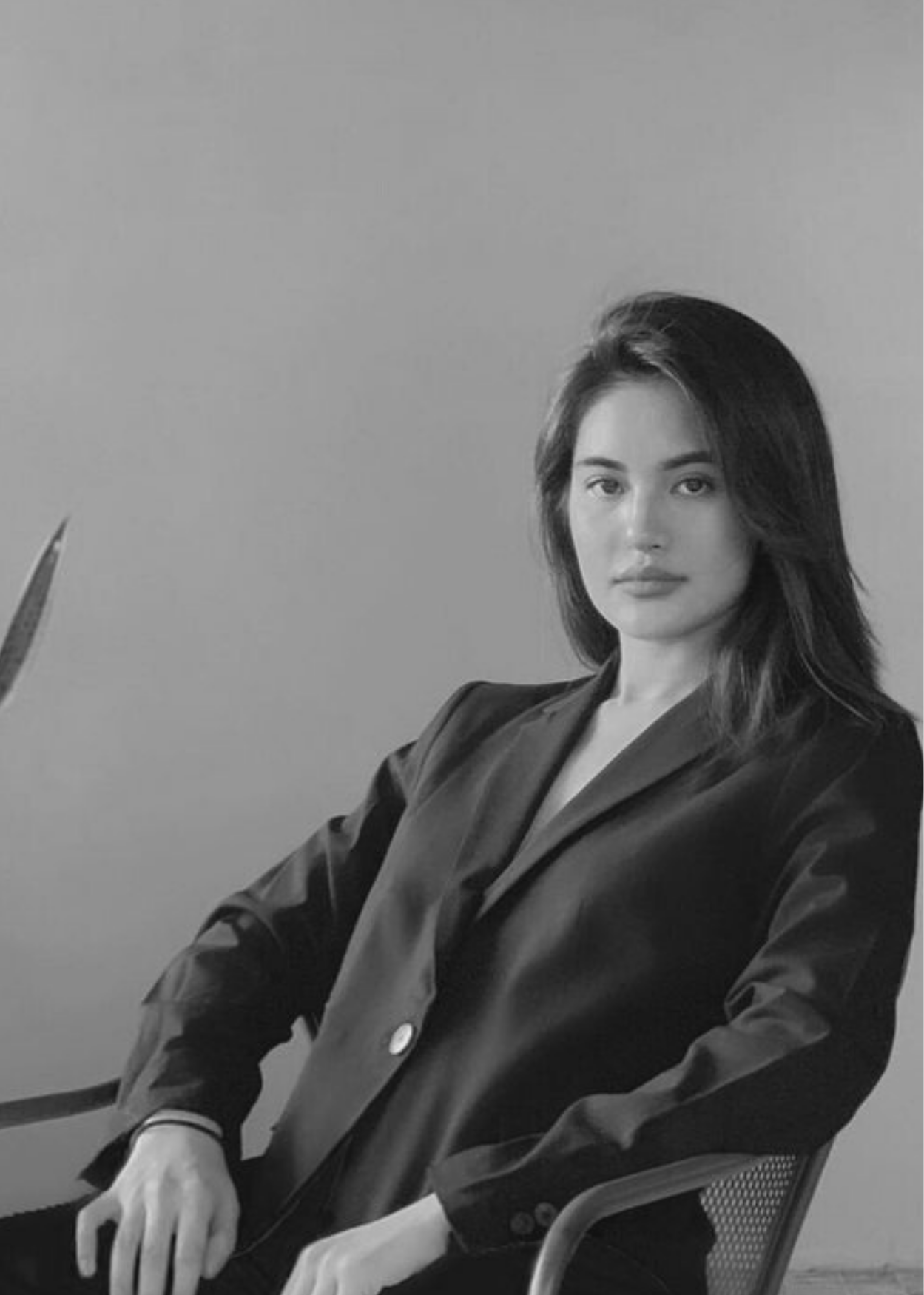 Confident woman with long hair wearing a dark blazer sitting on a chair against a plain background.