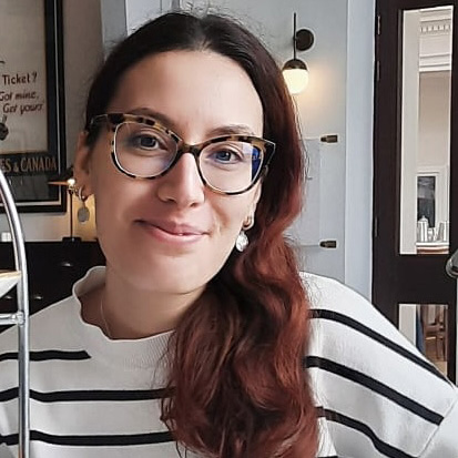 Woman with long reddish hair wearing tortoiseshell glasses and a white top with black stripes, smiling indoors.