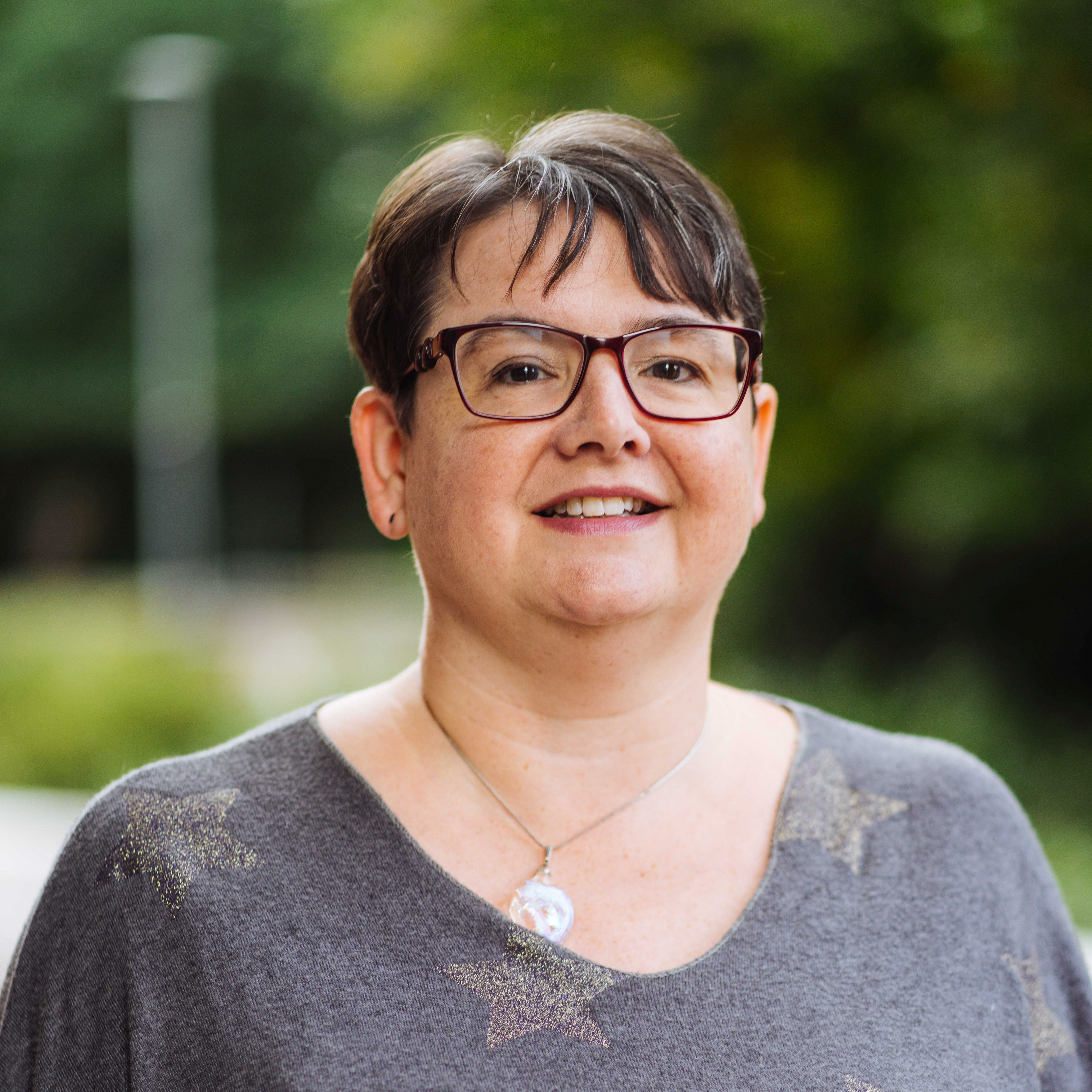 Smiling woman with short dark hair and glasses wearing a gray sweater, with a blurred outdoor background.