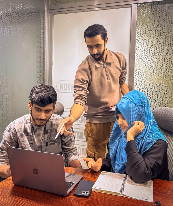 Three young adults working together at a table with a laptop, notebook, and smartphone, one person pointing at the laptop screen while another listens attentively.