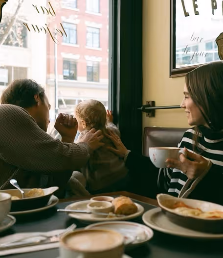 Drie mensen, waaronder een kind, zitten samen aan een cafétafel met koffie en gebak, terwijl ze naar buiten kijken.