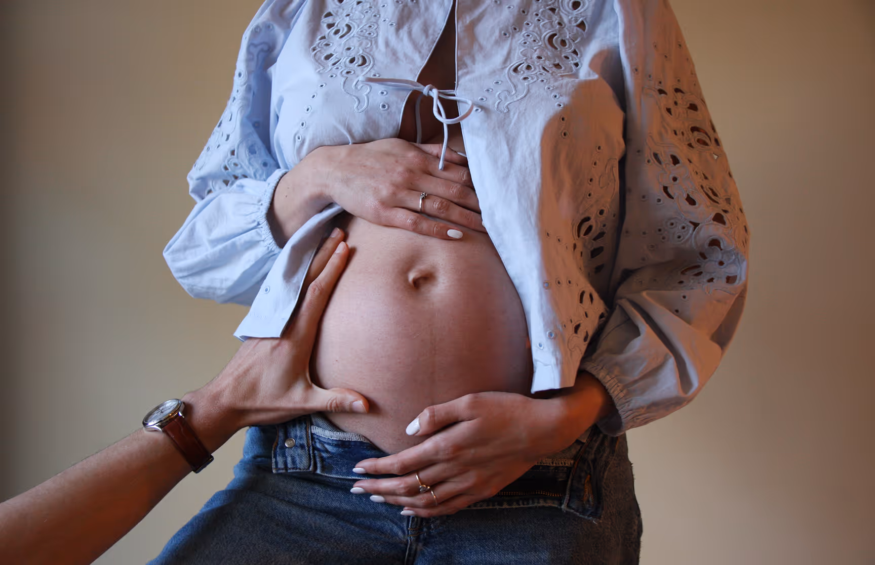 Handen van een man en vrouw die een zwangere buik vasthouden, vrouw draagt een lichtblouse en spijkerbroek.