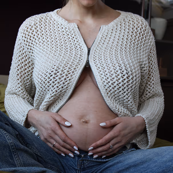 Zwangere vrouw in een witte gebreide trui en spijkerbroek, met haar handen liefdevol op haar blote buik.