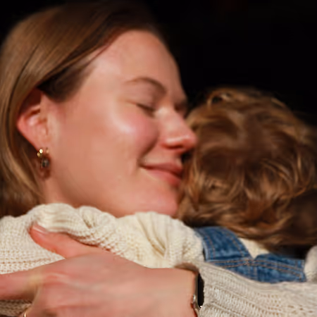 Vrouw met gesloten ogen en glimlach omhelst een kind met blond krullend haar en spijkerjasje.