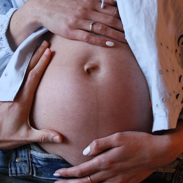 Buik van een zwangere vrouw die ondersteund wordt door handen met sieraden en witte nagellak.