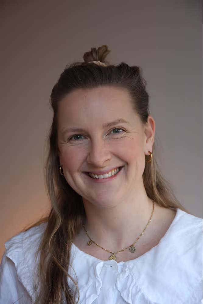 Vrouw met lang lichtbruin haar half opgestoken, glimlachend naar de camera, met een witte blouse en gouden sieraden.