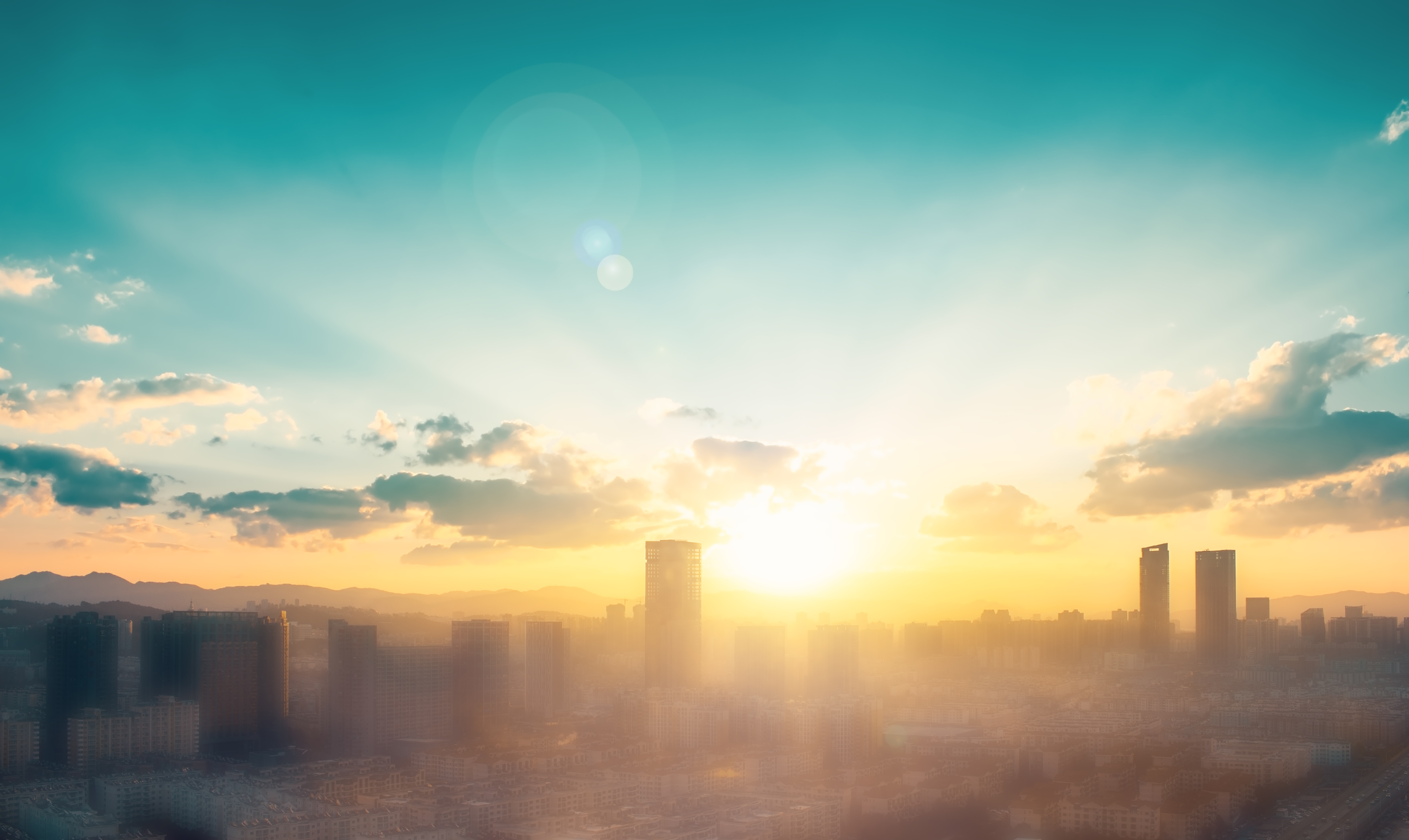 City skyline at sunrise with warm sunlight, scattered clouds and distant mountains, illustrating urban climate, heat and long-term climate change.