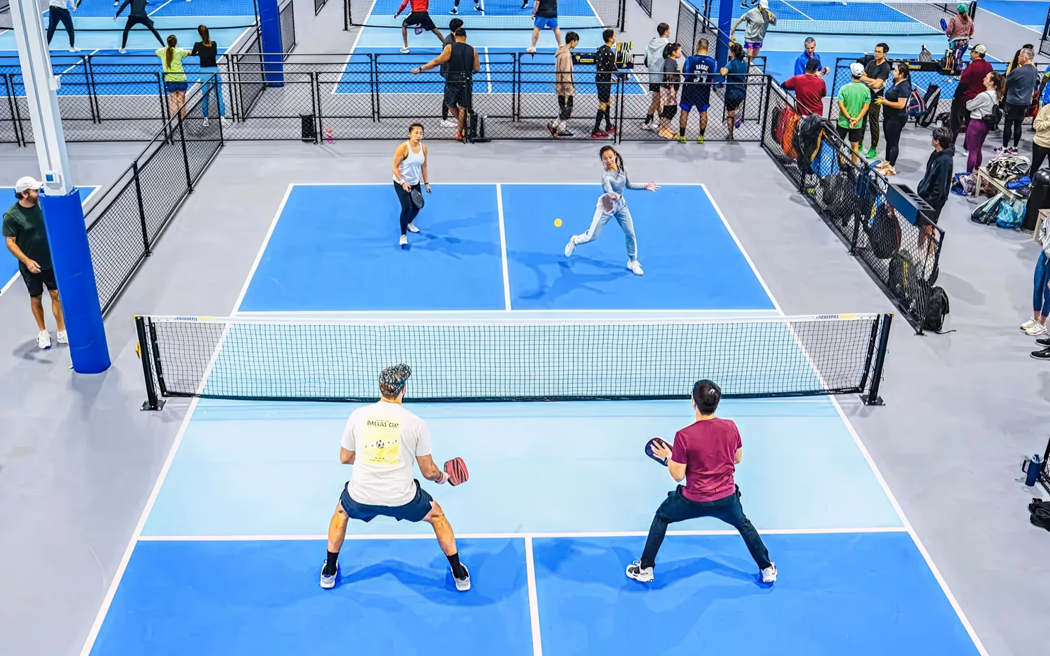 Four people playing doubles pickleball on an indoor blue court surrounded by spectators behind a fence.
