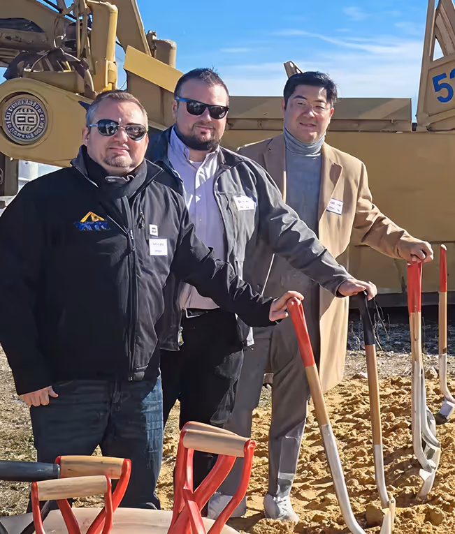 Three men wearing sunglasses and jackets stand side by side holding shovels at a groundbreaking ceremony with construction equipment in the background.