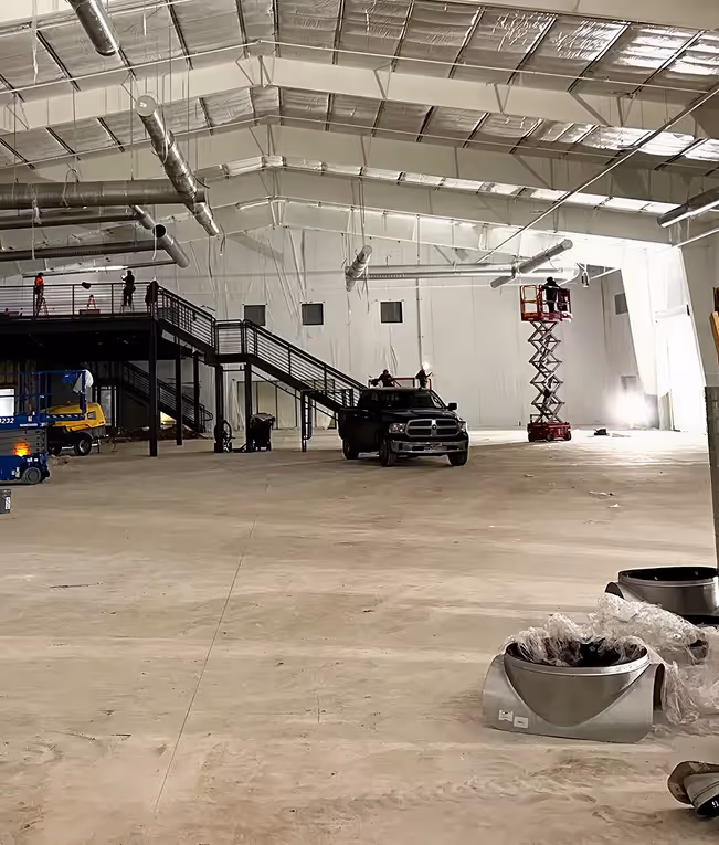 Large industrial warehouse interior with a concrete floor, metal staircase, black pickup truck, scissor lift, and overhead ductwork under construction.