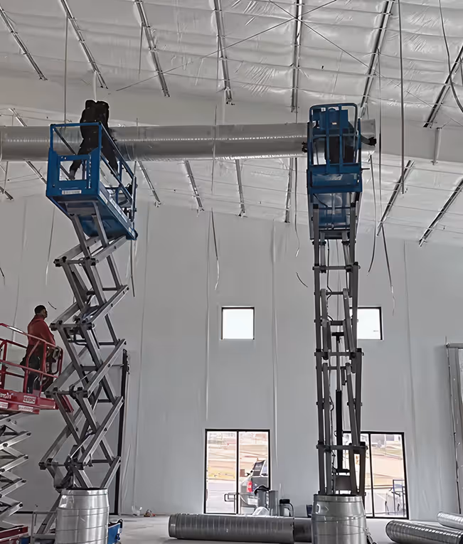 Two workers on blue scissor lifts installing ductwork inside a large industrial building with a high ceiling.