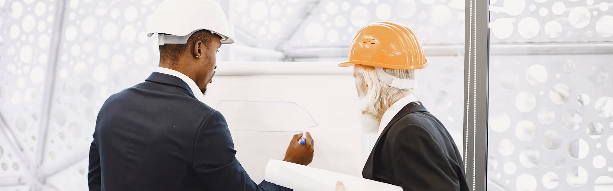 Two architects in suits and hard hats discussing a blueprint on a whiteboard.