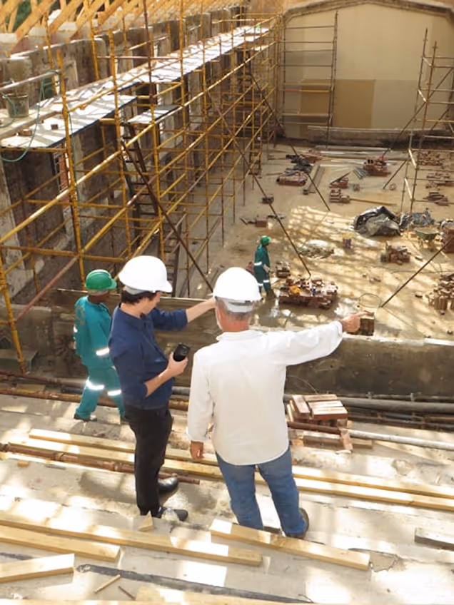 Two construction supervisors in white helmets inspecting a large building site with scaffolding and workers.