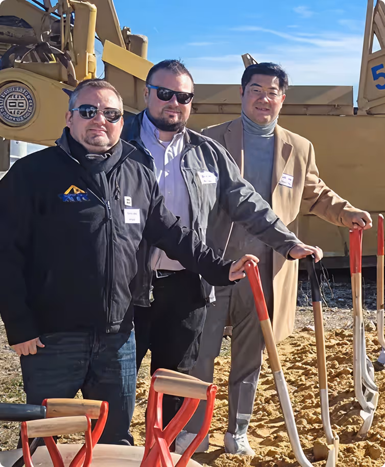 Three men standing outdoors holding shovels with a construction vehicle in the background.