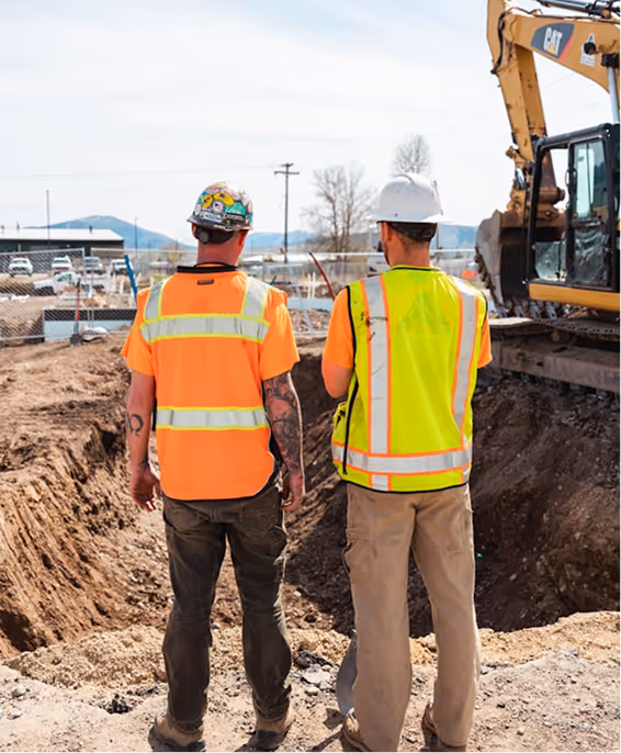 Two construction workers in reflective vests and hard hats standing by a deep excavation site with an excavator nearby.