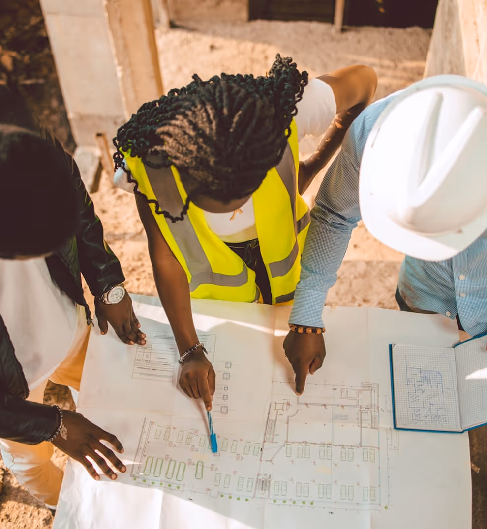 Three construction professionals reviewing blueprints on a table at a worksite, one wearing a yellow safety vest and another a white hard hat.