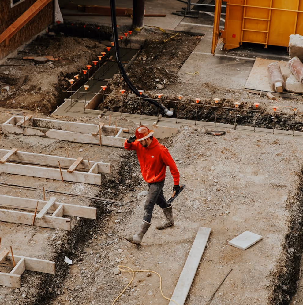 Construction worker in a red hoodie and hard hat walking across a dirt site with wooden framing and rebar.