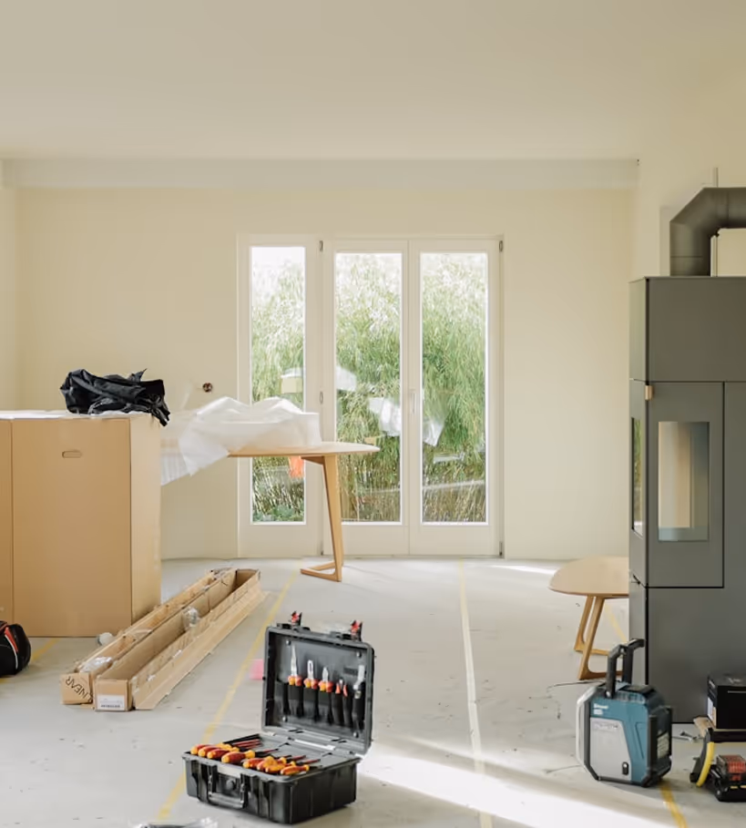 Bright, minimalistic room under renovation with tools in an open black case on the floor, large windows with greenery outside, and a modern wood stove on the right.